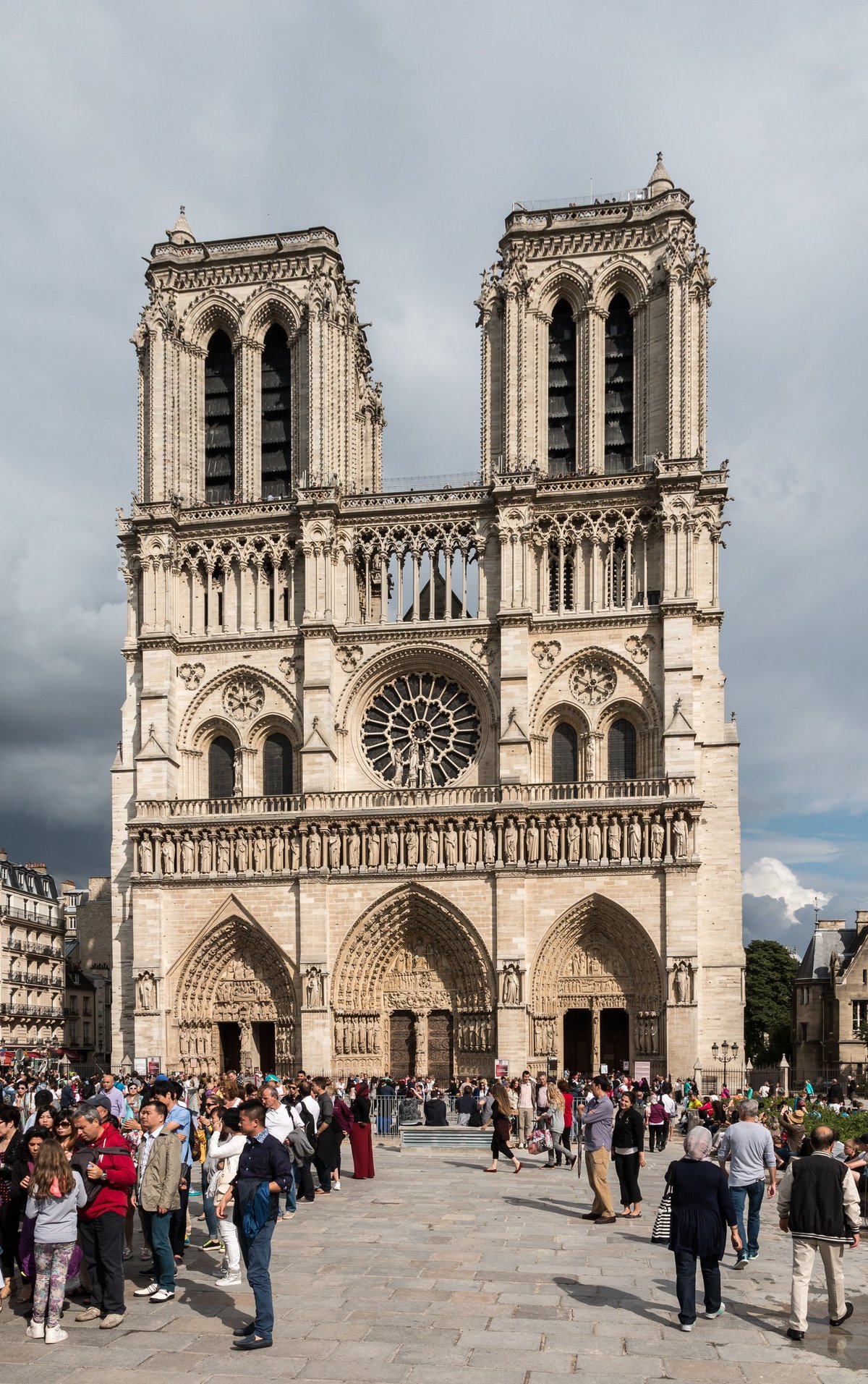 The west front of Notre-Dame de Paris rising over a busy parvis under a dramatic sky